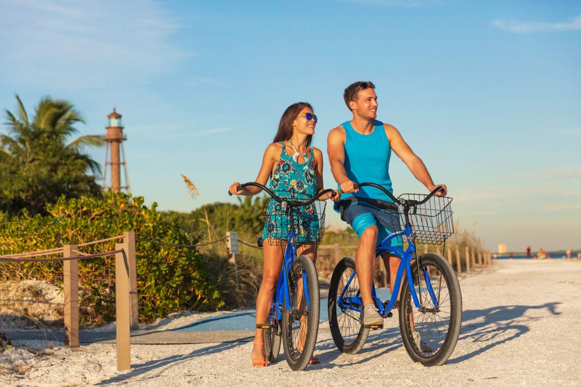 Florida beach vacation couple biking sport rental bikes recreational activity happy watching sunset on Sanibel Island by the Lighthouse. Young woman and man riding bicycles. Summer people lifestyle .